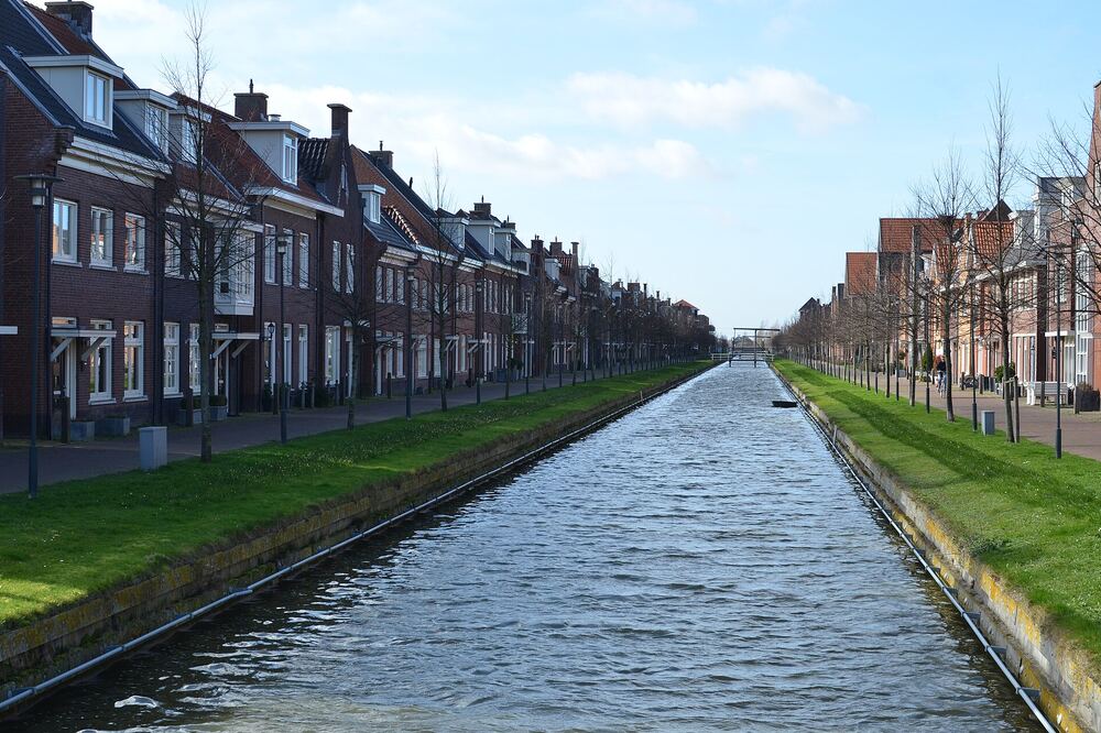 Rechte gracht midden in Berkel en Rodenrijs met aan beide zijden rijen bakstenen woningen, jonge bomen en grasbermen, en in de verte een ophaalbrug onder een blauwe lucht